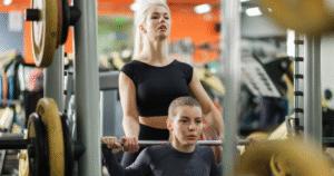 two females working out in a gym together. one lifts while her workout buddy spots her.